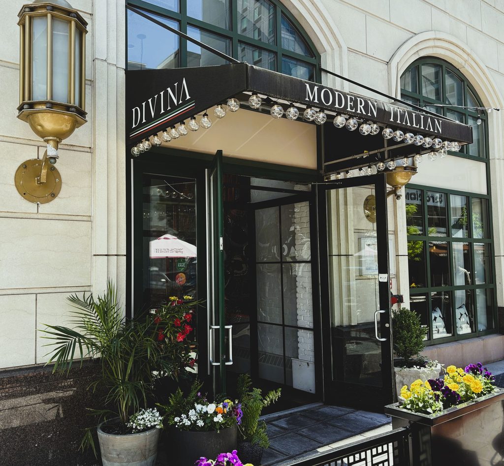 Exterior storefront of Divina Modern Italian restaurant featuring a black lighted awning, open glass double doors, and outdoor potted plants.