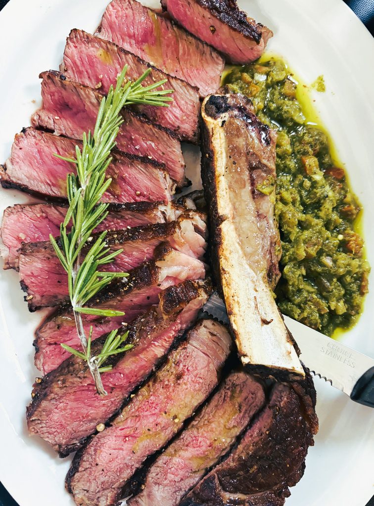 Overhead view of a sliced, medium-rare steak served next to a large bone, garnished with a rosemary sprig and a side of green chimichurri sauce.