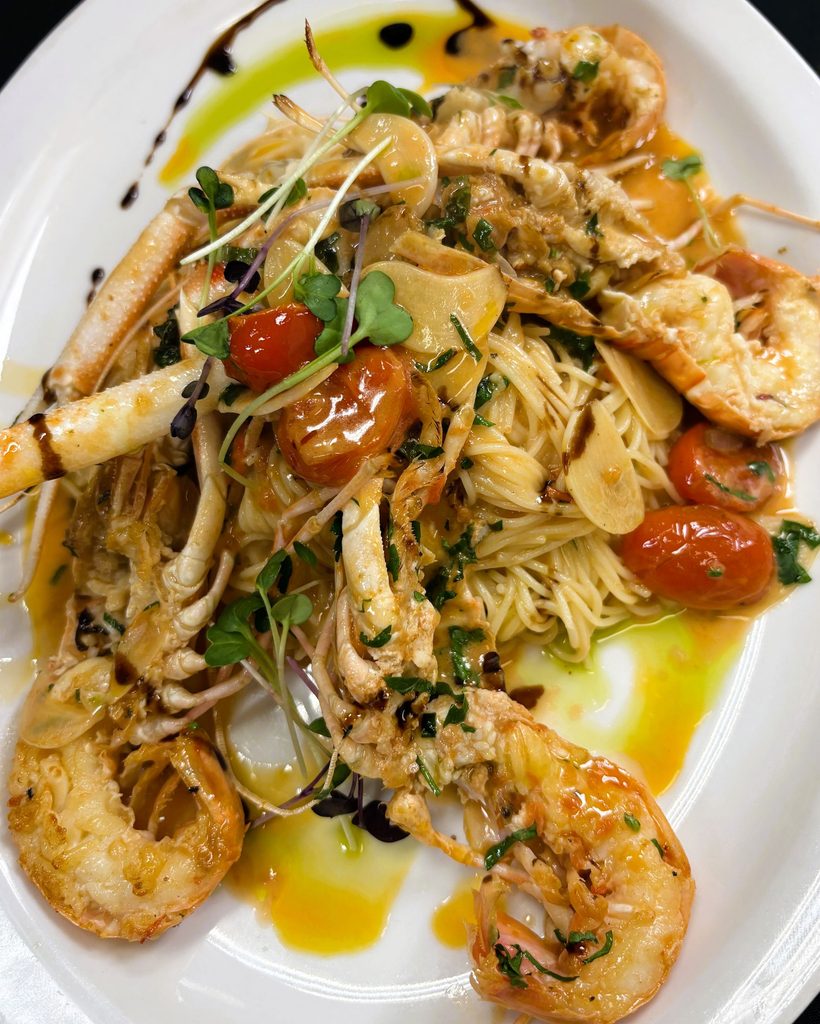 Seafood pasta dish plated with large shrimp, roasted cherry tomatoes, garlic, and fresh microgreens.