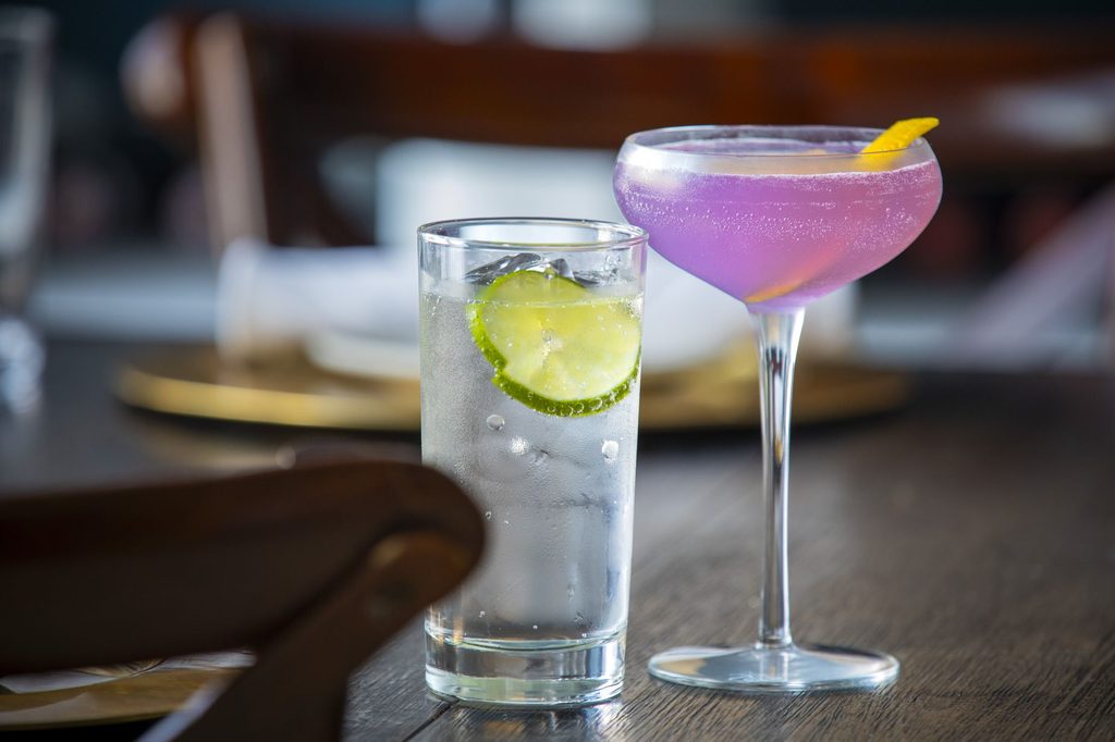 Two cocktails on a bar table: a tall, iced clear drink with a lime wheel, and a bright purple drink served in a coupe glass with a lemon twist.