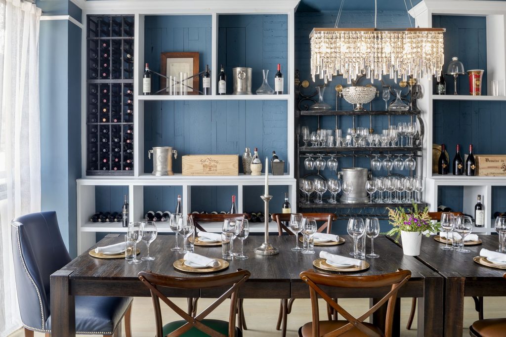 Bright dining room interior featuring a long set table with wooden cross-back chairs and large blue built-in shelves displaying wine and glassware.