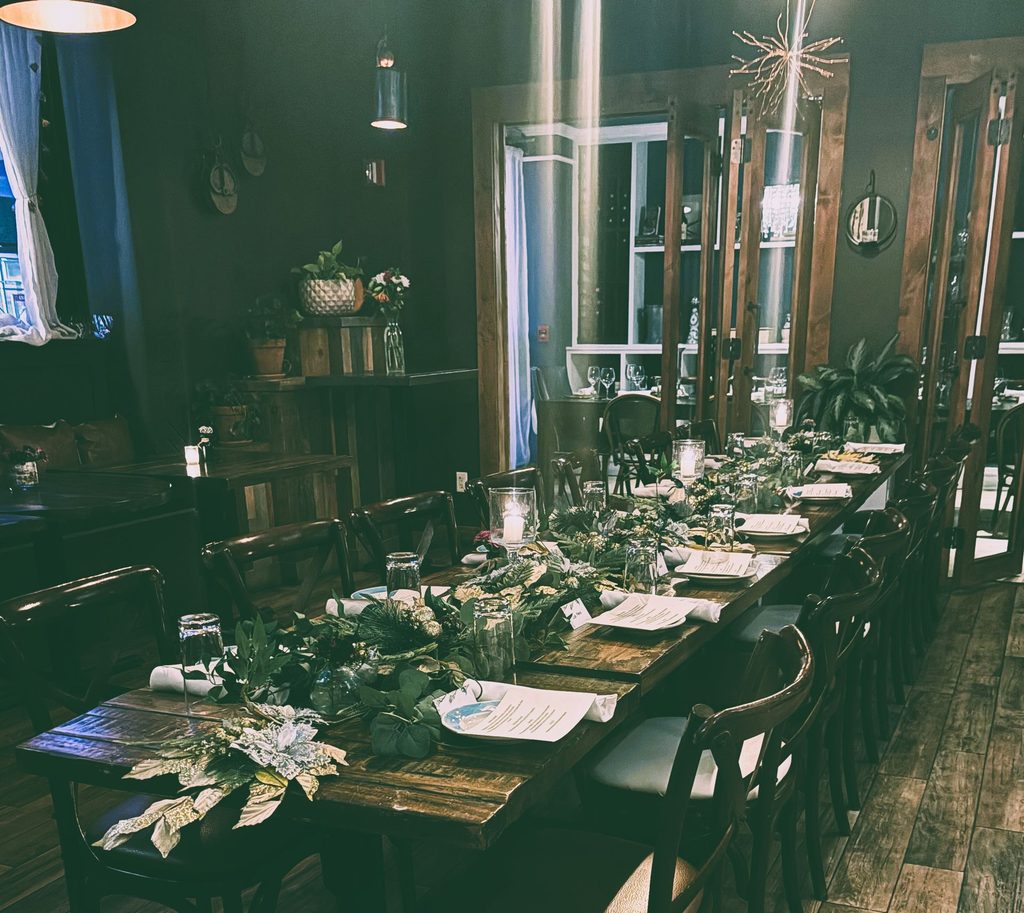 Intimate, softly lit dining room featuring a long rustic wooden table set for an event with a lush green garland runner and glassware.