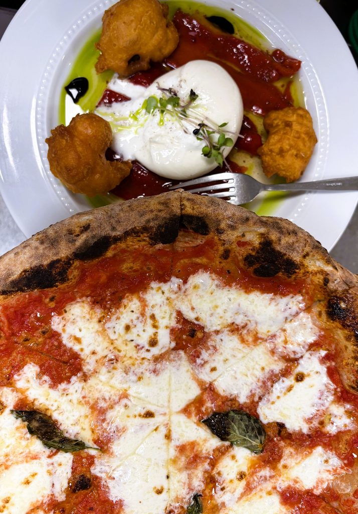 A wood-fired Margherita pizza shown alongside an appetizer plate of creamy burrata, roasted red peppers, and fried dough.