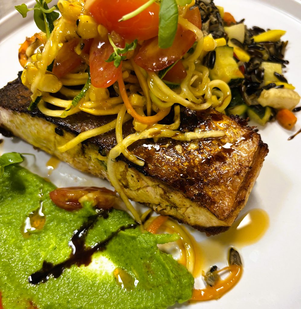 Close-up of a thick, seared fish fillet topped with a fresh tomato and squash salad, resting on a bright green puree with balsamic drizzle.