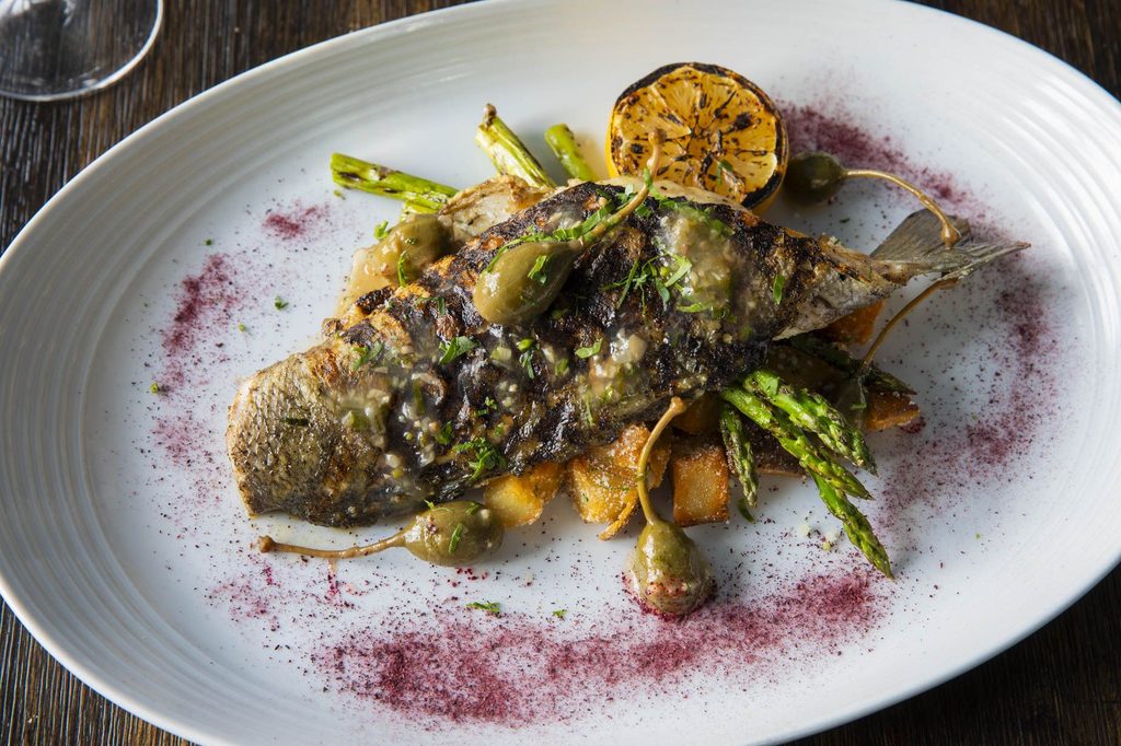 Whole grilled fish served over roasted potatoes and asparagus, garnished with capers, herbs, and a charred lemon half.
