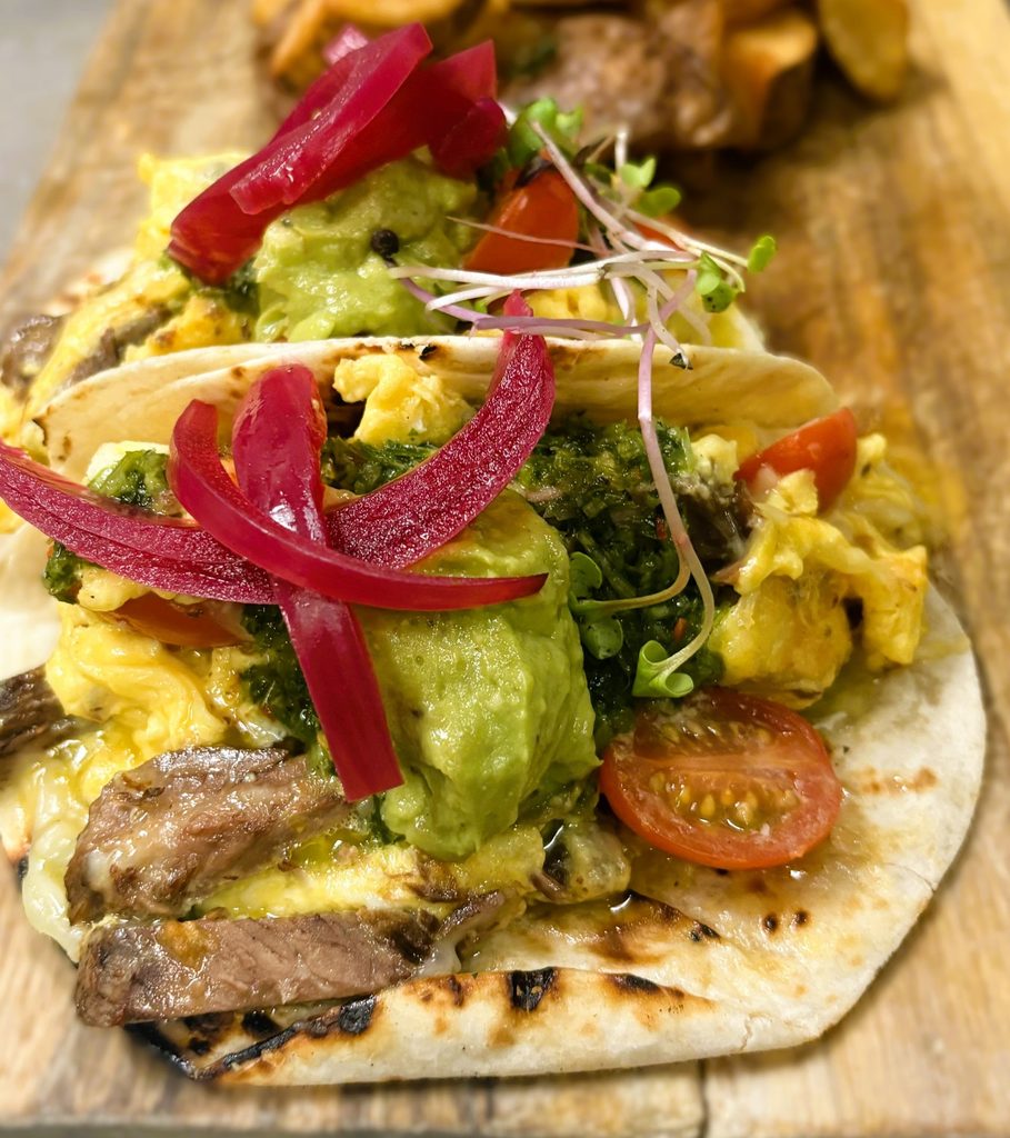 Two breakfast tacos on a wooden board filled with scrambled eggs, steak, guacamole, and bright pink pickled red onions.