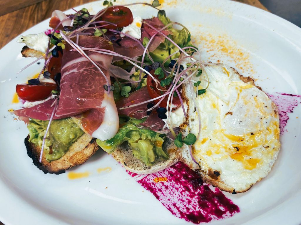 Breakfast plate featuring avocado toast topped with sliced prosciutto, a sunny-side-up fried egg, and microgreens.