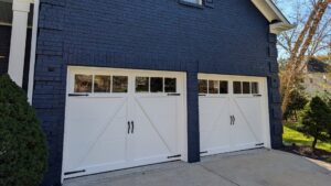 Two single white carriage-house garage doors with black decorative hardware and diagonal bracing on a dark navy brick exterior.