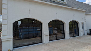Three custom arched full-view glass garage doors with black aluminum frames installed in white stucco archways.