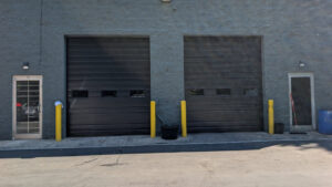 Two heavy-duty black commercial sectional garage doors with small rectangular vision lites on a grey block industrial building.