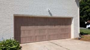 Double-wide faux wood garage door with a natural oak finish and long flush panels on a painted white brick exterior.