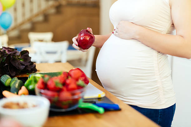 a pregnant woman eating an apple standing at a table with healthy foods like fruits and vegetables, promoting a healthy pregnancy